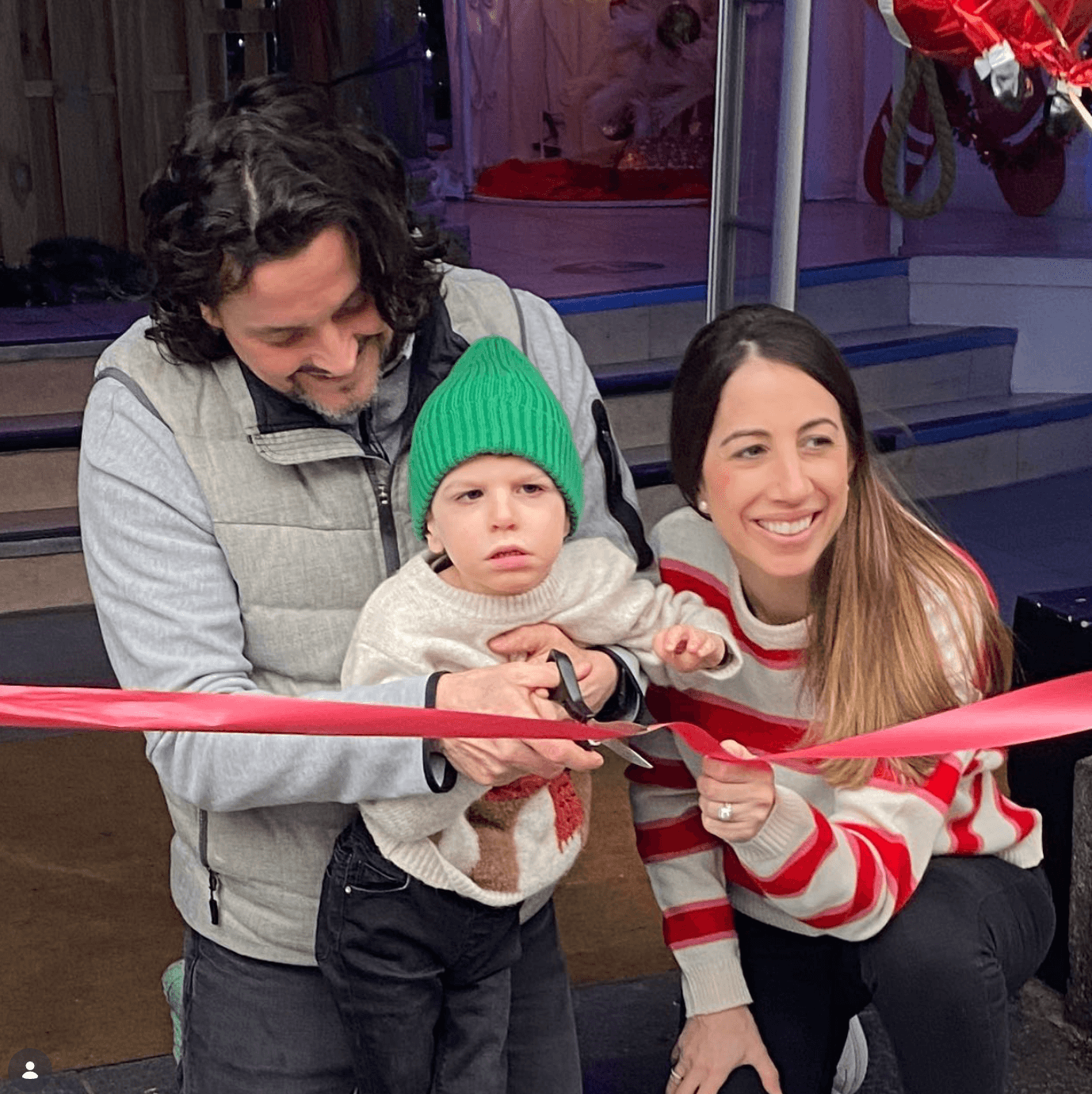 Opening day of the first grotto: a child in a green beanie cuts a red ribbon with help from a parent while the other parent smiles beside them.